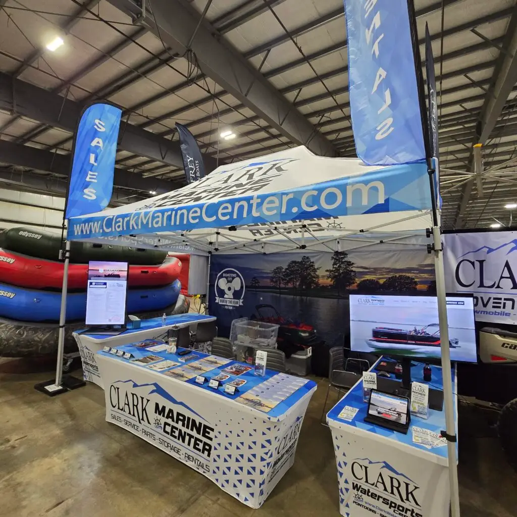Durable-Trade-Show-Marketing-Materials - 2X Sales Trade show booth for Clark Marine Center with blue sale flags, banners, and stacked inflatable boats on display in an indoor expo hall.