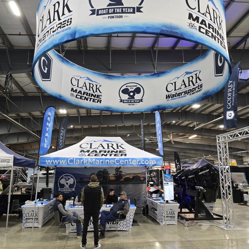 custom-Trade-Show-Marketing-Materials - 2X Sales Large circular blue-and-white booth banner hanging from the ceiling advertising Clark Marine Center at a boat show, with people seated at tables underneath.